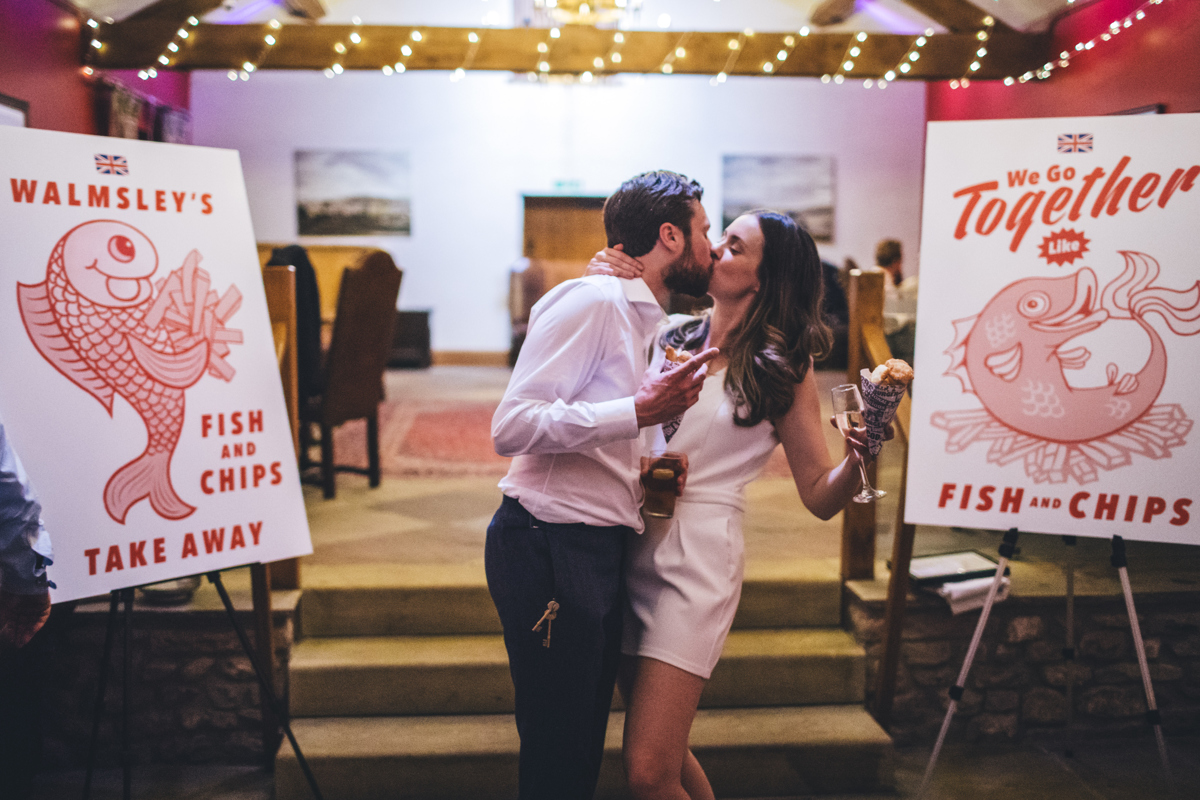 Bride and groom kissing next to fish and chip posters at their wedding reception
