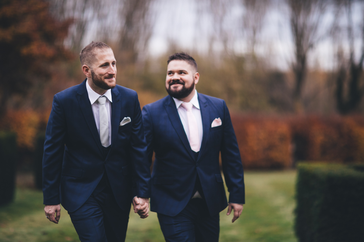 Two grooms holding hands walking through formal gardens
