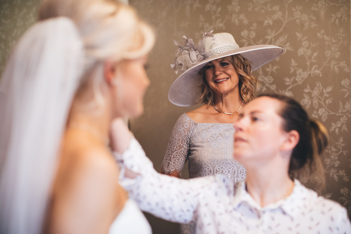 Mother of the bride in the background looking on at the bride in the foreground who is having final touches done to her makeup
