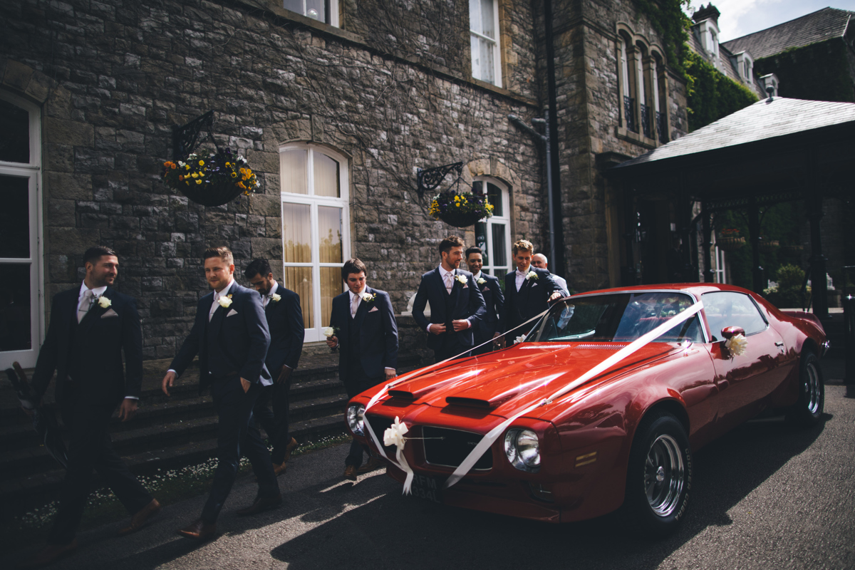 Groom and groomsmen walking past a bright red Pontiac car used as the wedding car