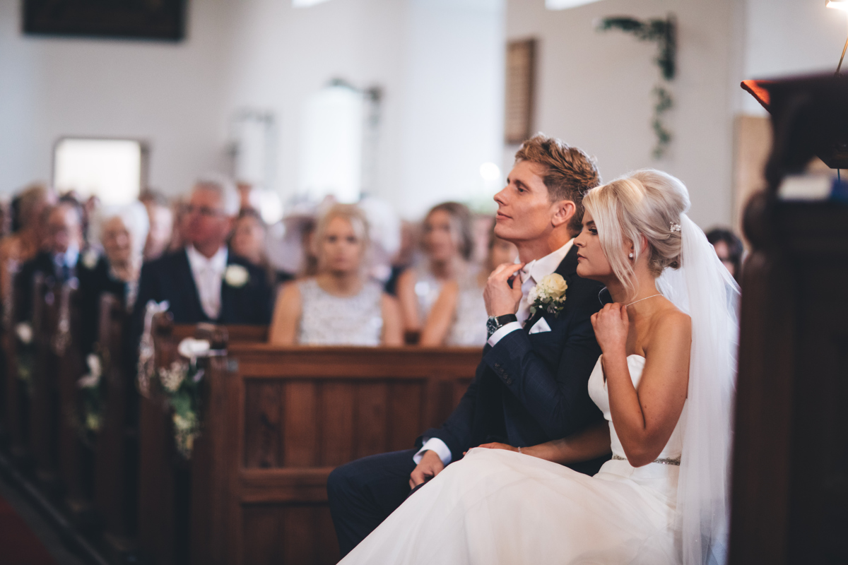 Bride and Groom sat together at the front of the church, both of them are touching their necks