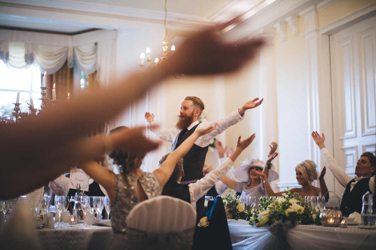 Wedding party seated with their arms in the air with one man stood also with his arms in the air