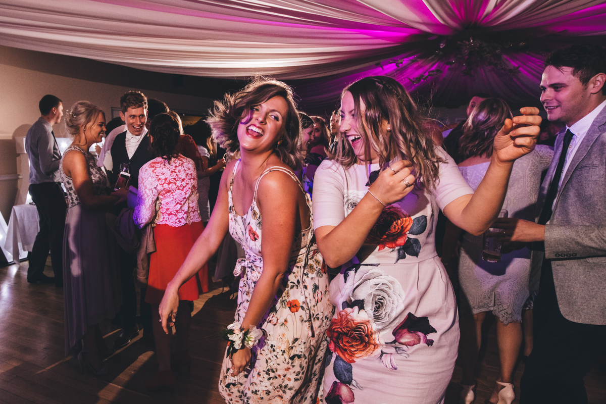 Two female wedding guests in floral print dresses laughing and dancing with their backs to one another