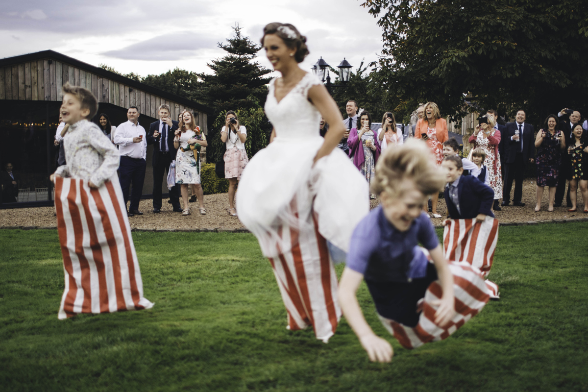 Bride and children jumping in a sack race with the wedding party laughing in the background