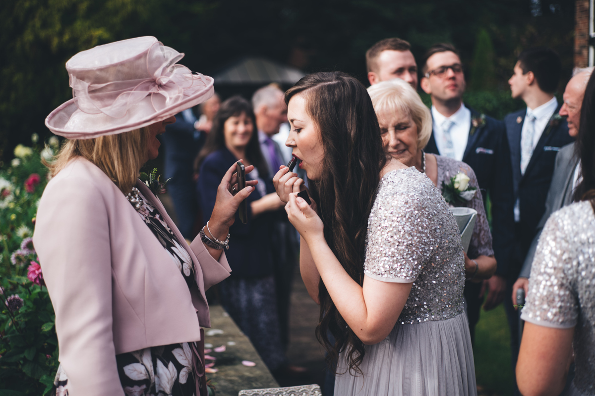 Bridesmaid applying lipstick with the help of a guest holding a mirror