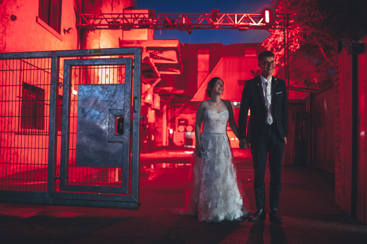 Bride and Groom holding hands standing next to a large metal gate backlit with red light