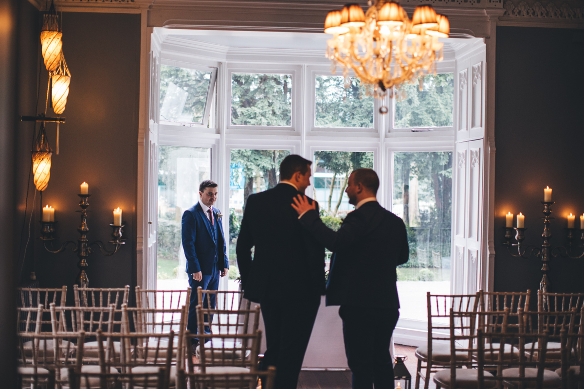 Groom in the bay window facing two groomsmen who are walking down the aisle towards him