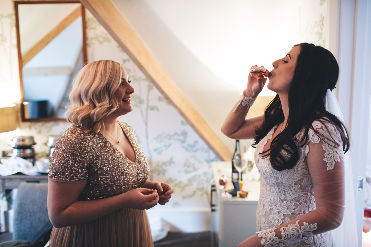 Bride having a quick drink to calm the nerves whilst one of her bridesmaids is stood opposite her smiling