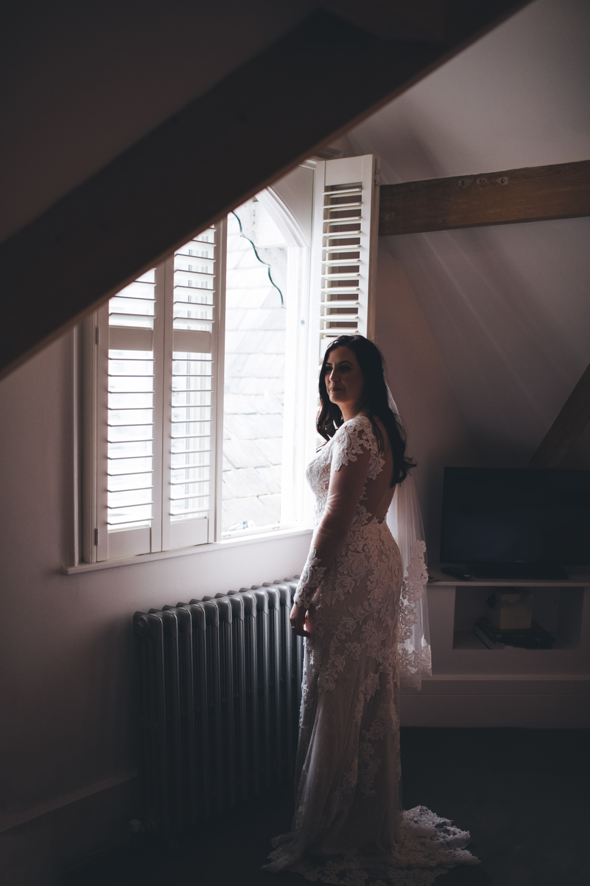 Bride stood in her wedding dress in front of a window with shutter blinds with a cast iron radiator below