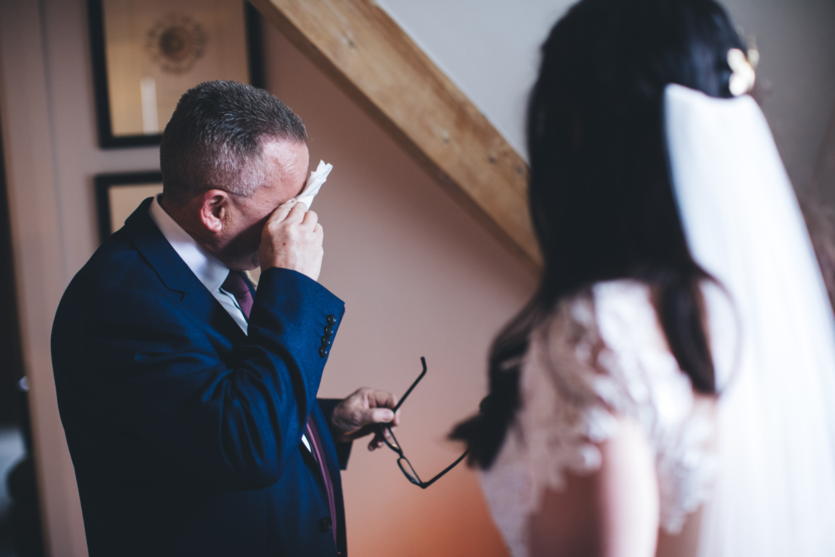 Father of the bride wiping away tears with a tissue and holding his glasses with his other hand with the bride in the foreground facing him