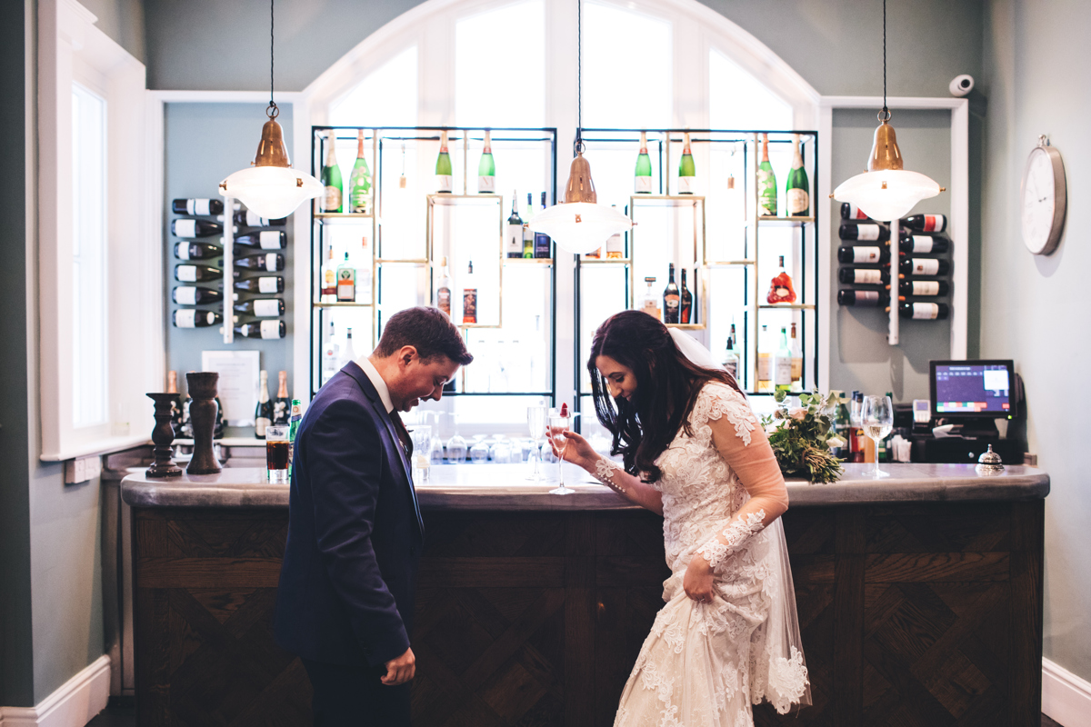 Bride and groom stood opposite each other at a bar looking down at the floor. The bride is holding a champagne glass