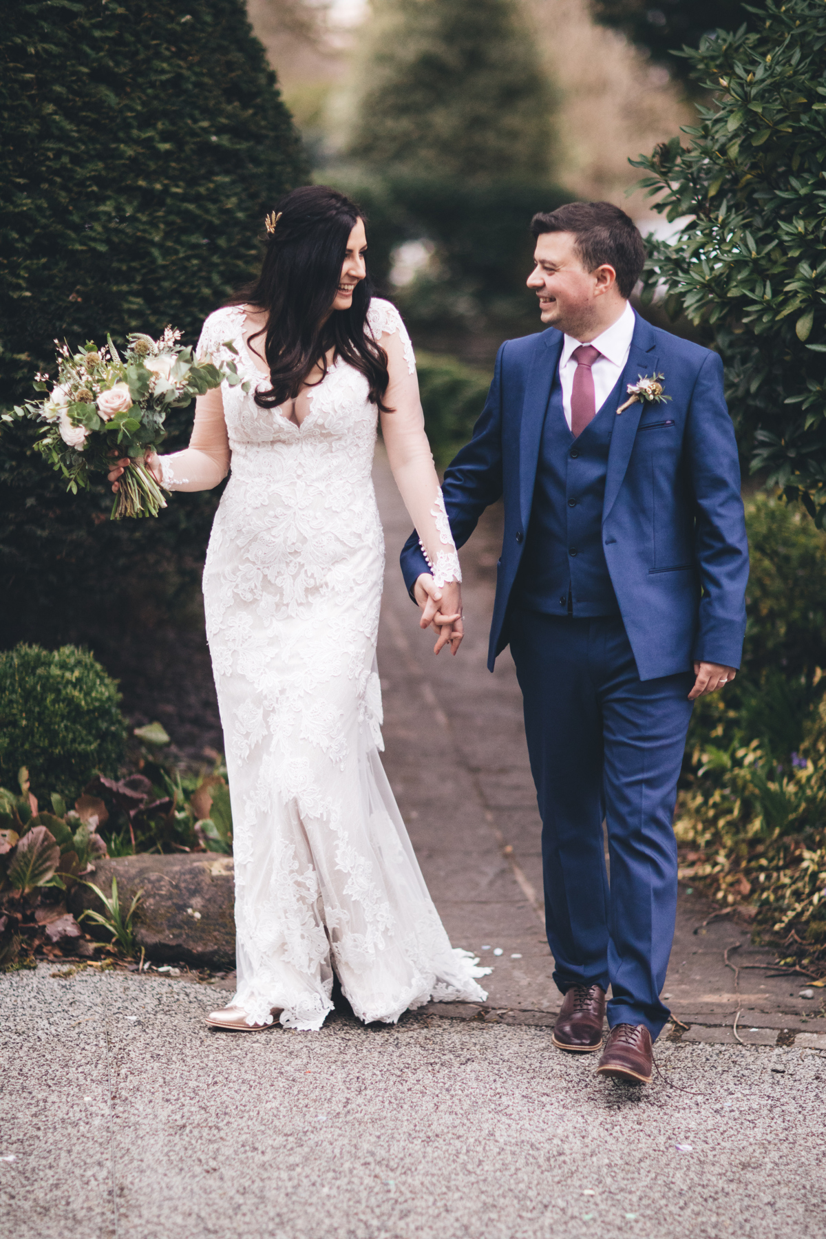 Bride and groom holding hands walking down a garden path with greenery on either side of them