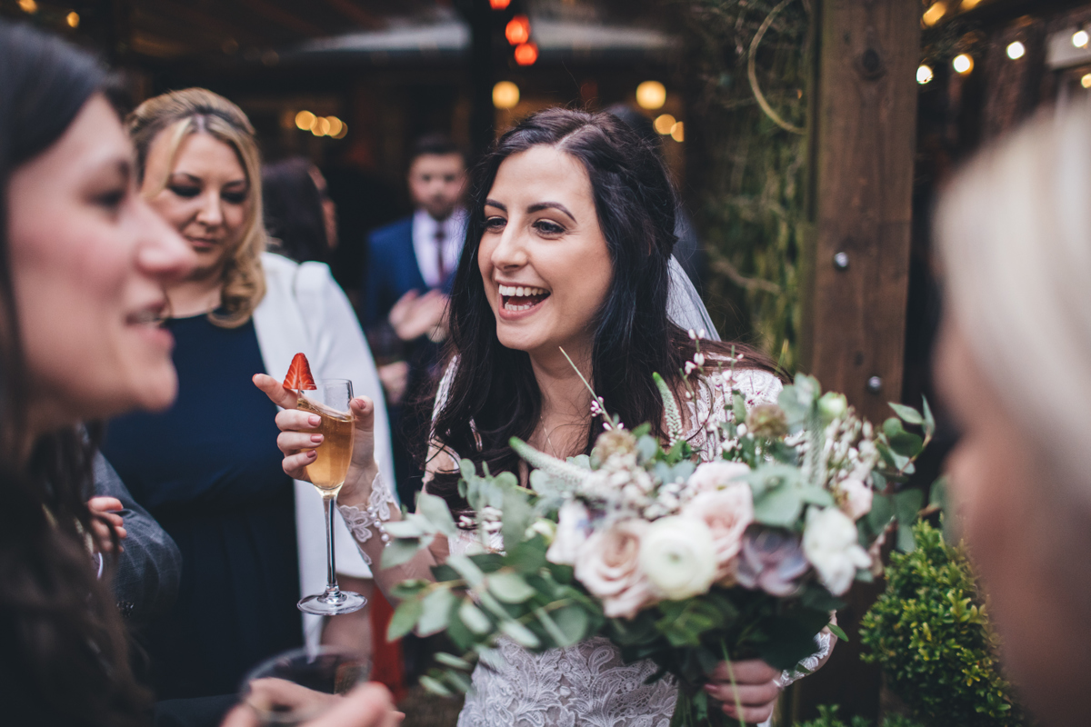 Bride smiling with a glass of champagne in one hand and her bouquet in the other chatting to her guests