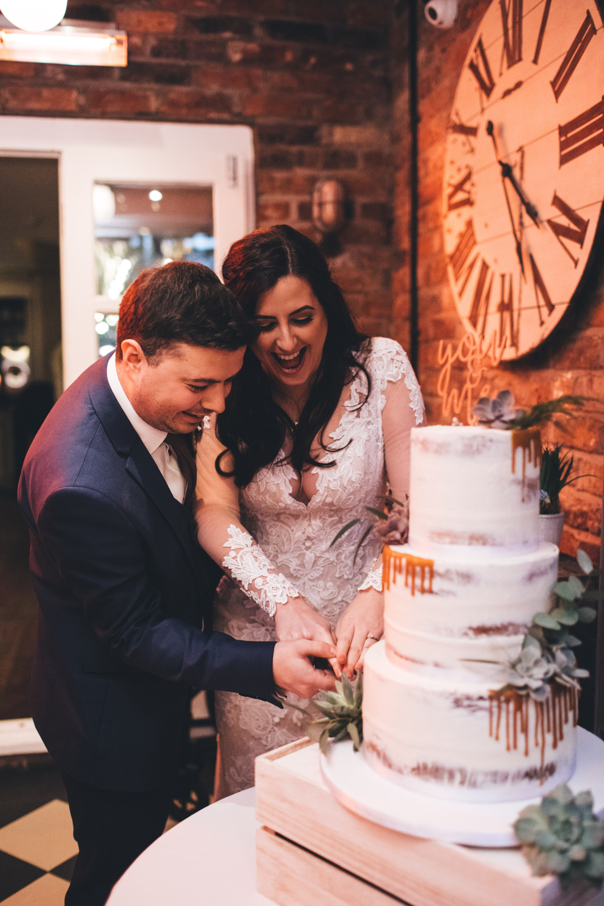 Bride and Groom cutting their three tier wedding cake. There is a large clock with roman numerals on the brick wall above the cake