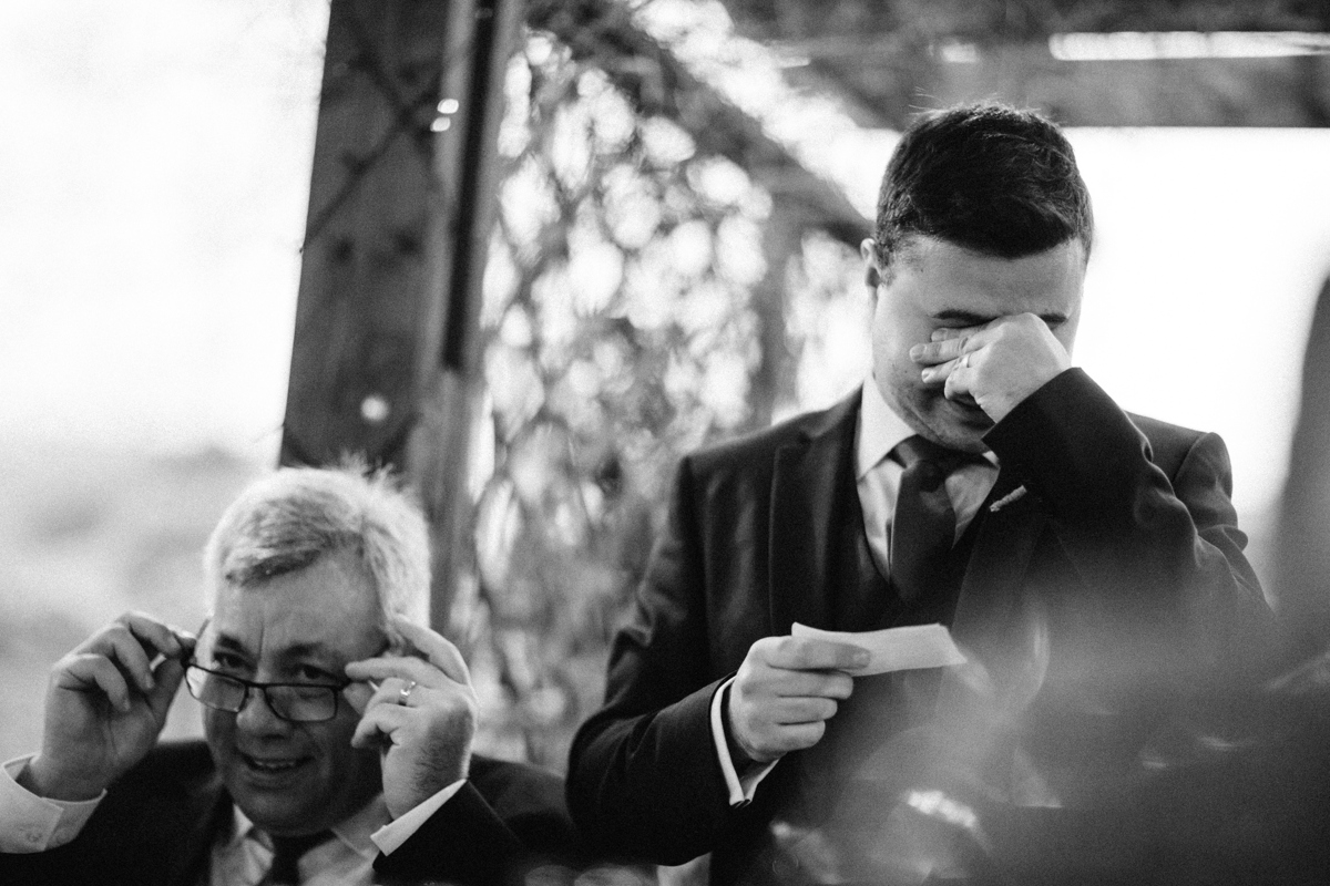 Black and white photograph of the groom stood doing a speech with his hand over his eyes whilst his father is sat next to him adjusting his glasses