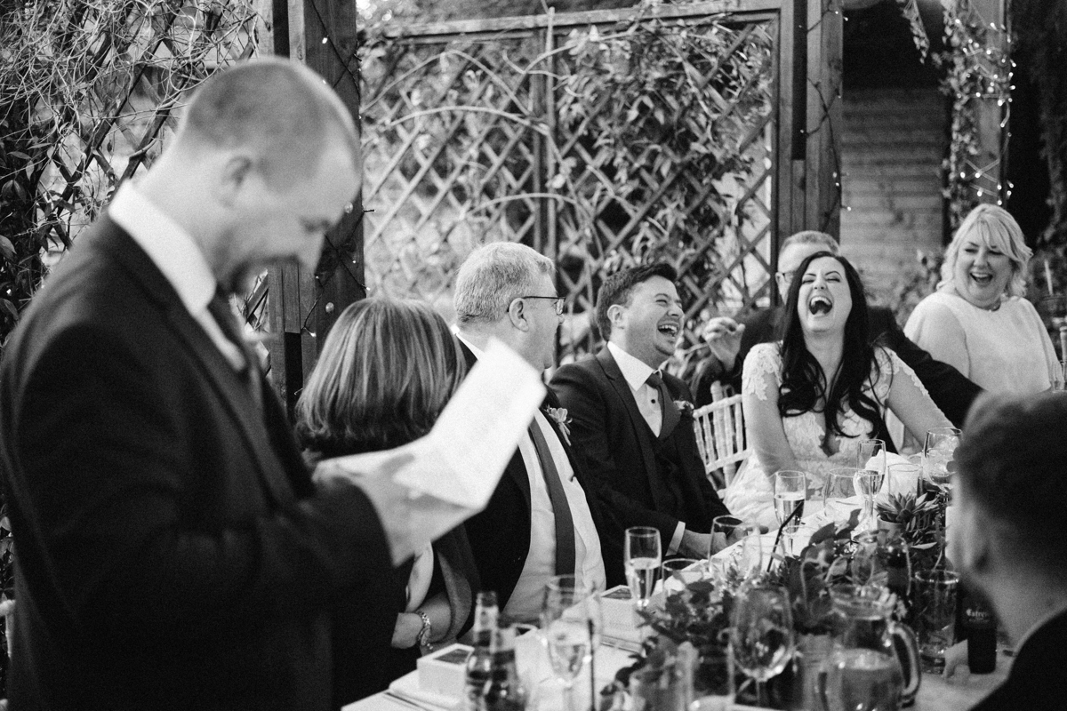 Best man stood reading his speech in the foregroud with the couple and their parents sat laughing