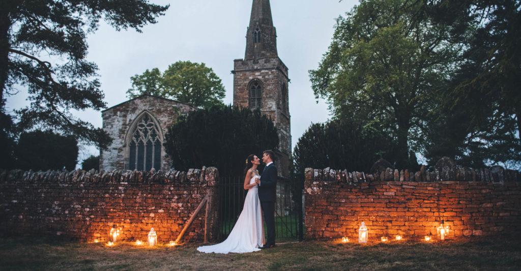 Bride and groom stood in front of a gate in an old stone wall which is lit by lanterns from below. There is a church in the background