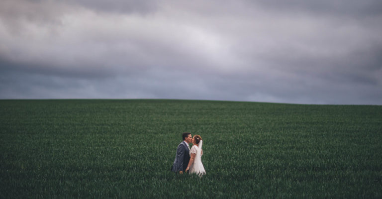 Bride and groom stood kissing in a large green field with green leaves coming up to their waists. Behind is a cloudy sky which looks like it might rain