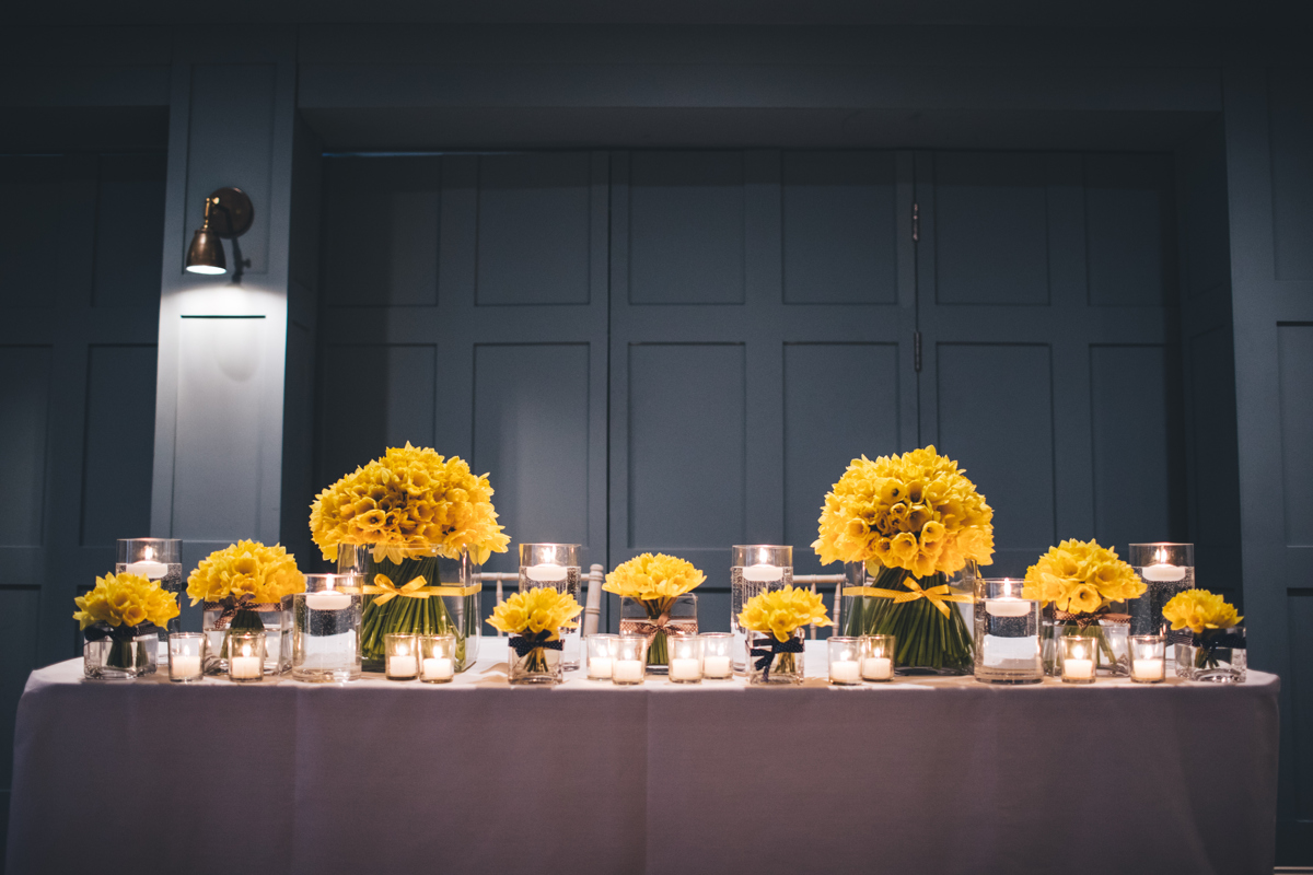 Daffodil arrangements in vases interspersed with candles on a large table covered with a white cloth