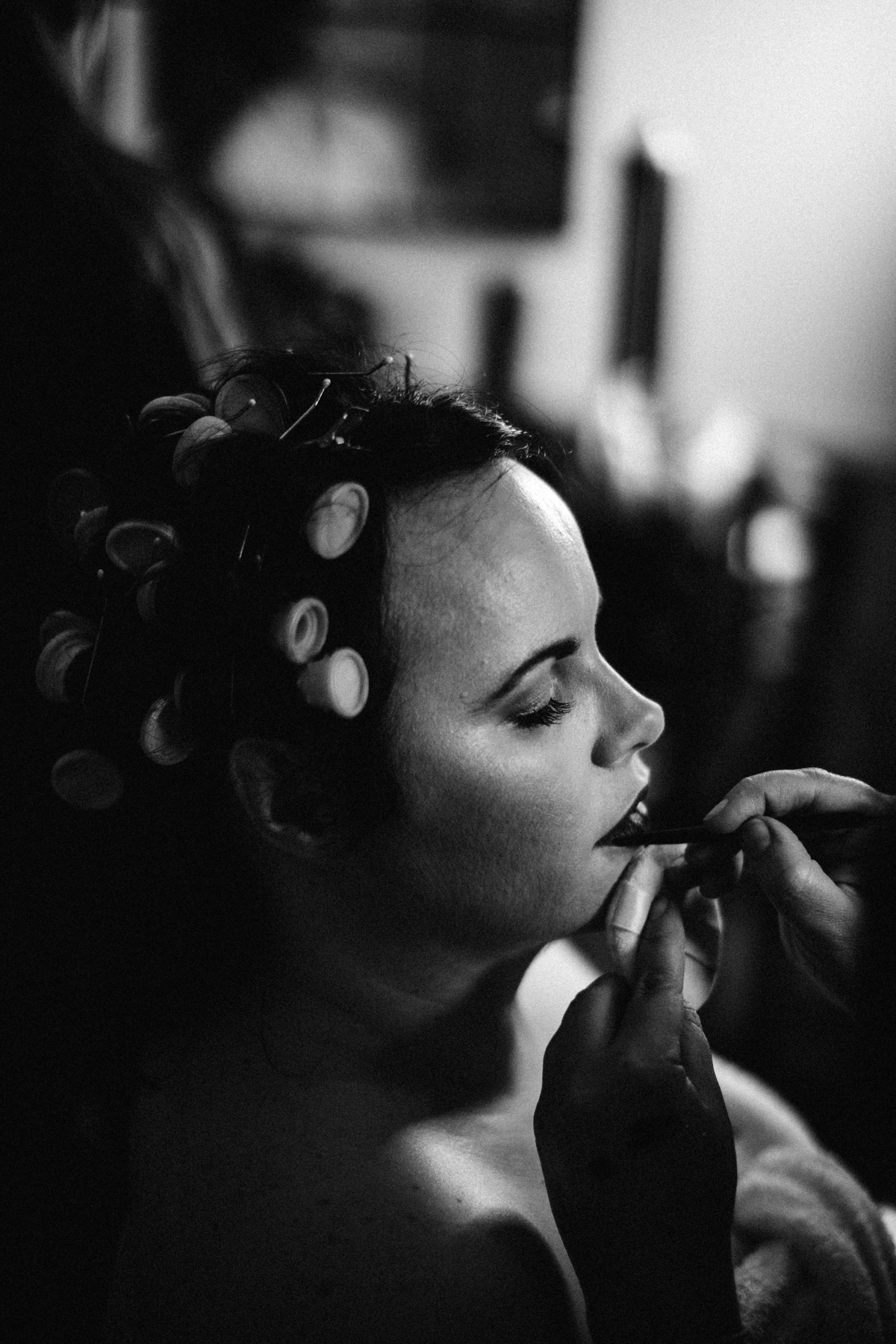 Black and white photograph of bride having her makeup done with rollers in her hair