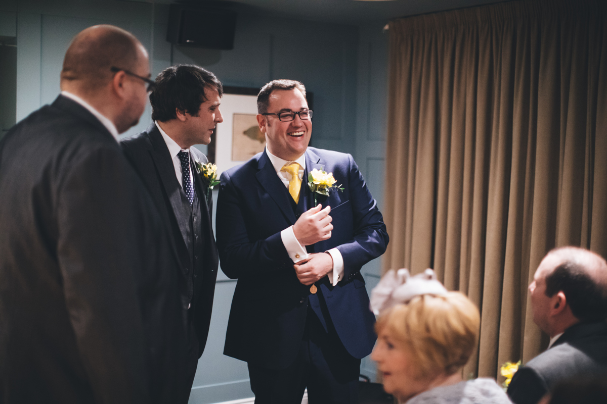 Groom and groomsmen stood waiting at the front of the room for the bride. There is a large beige curtain to the right of the picture