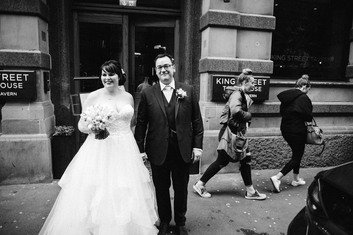 Black and white photograph of bride and groom stood holding hands outside King Street Townhouse, Manchester whilst two oung women walk past in the background