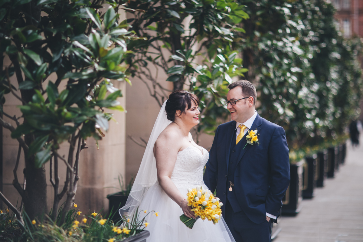Bride holding a daffodil bouquet stood next to her husband in front of tall green plants
