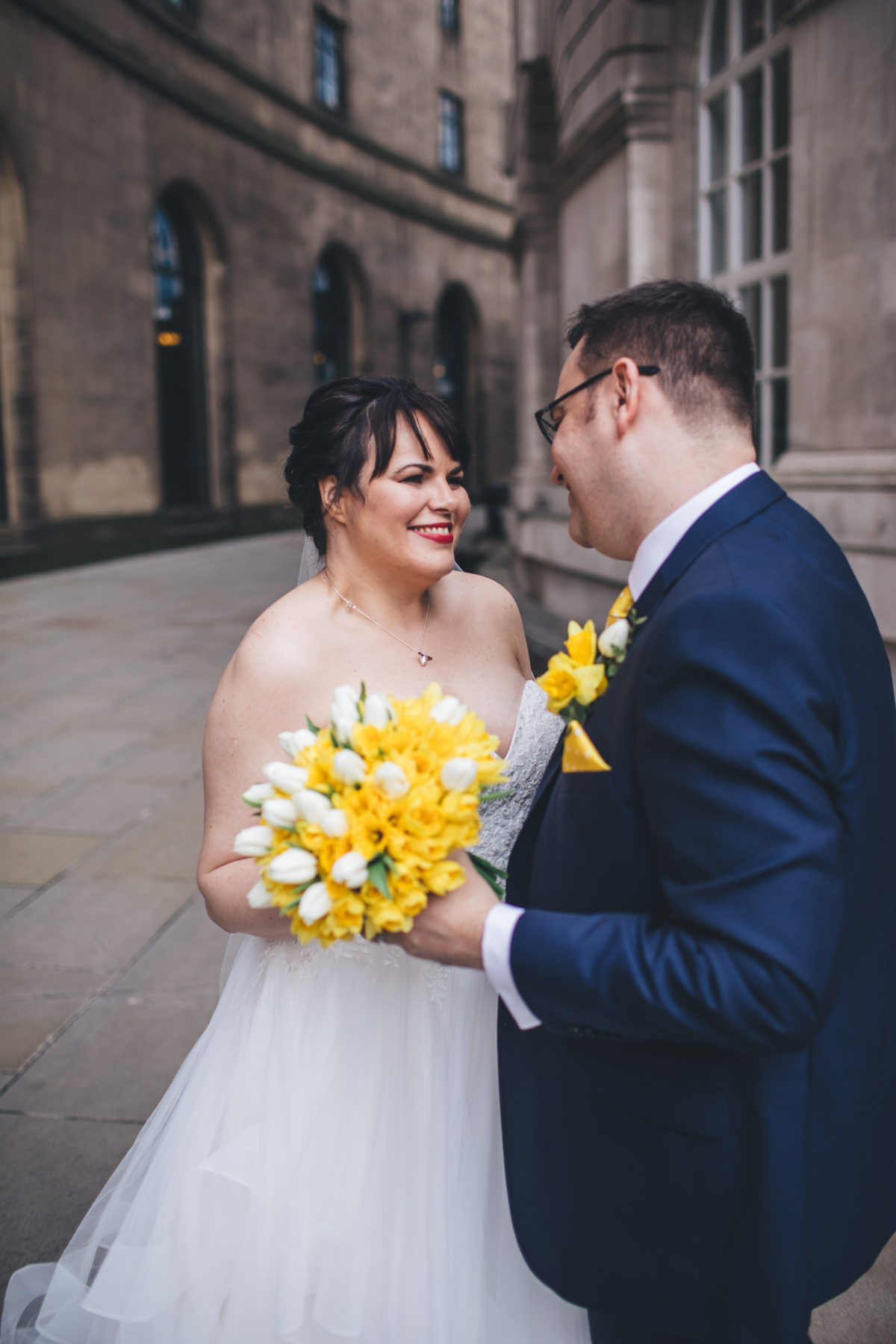 Bride and groom stood opposite each other smiling holding a bouquet of Daffodils between them