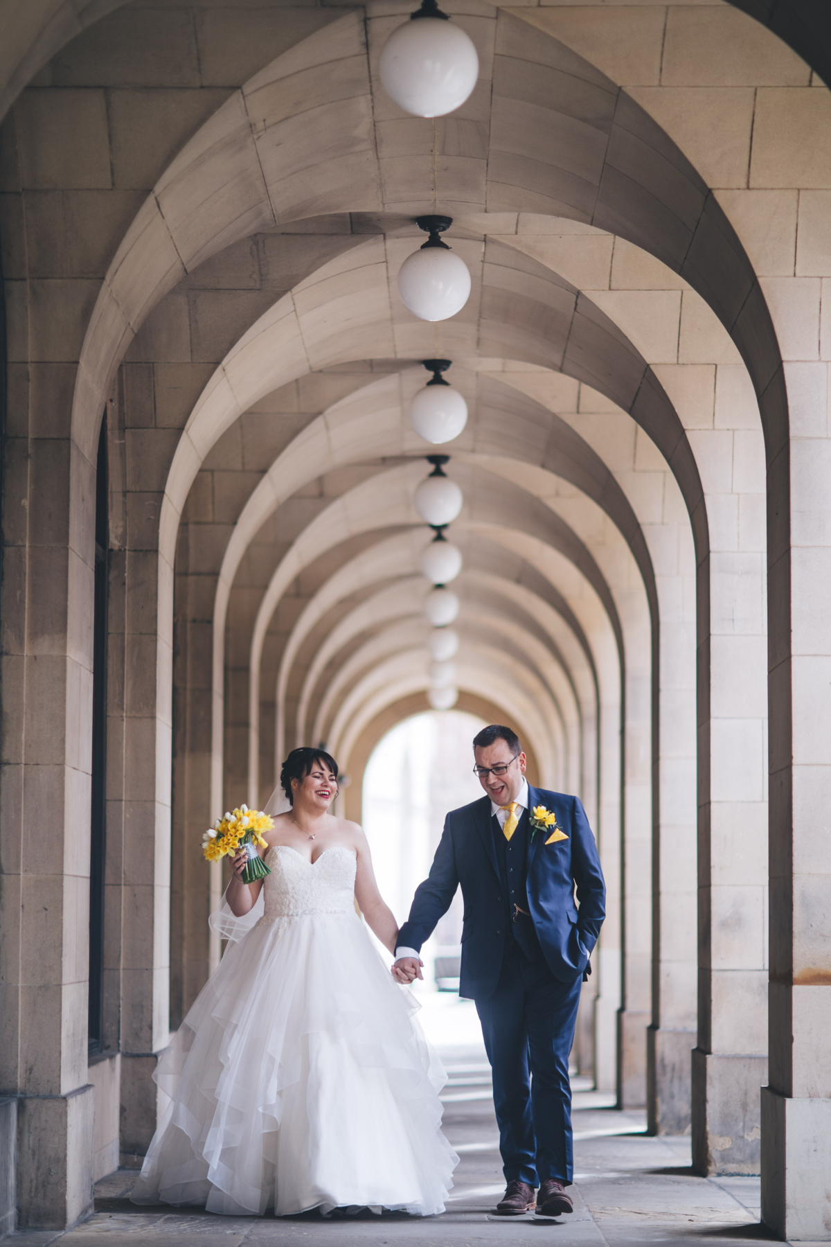 Bride and groom walking hand in hand along a outdoor corridor of arches with circular lights above them