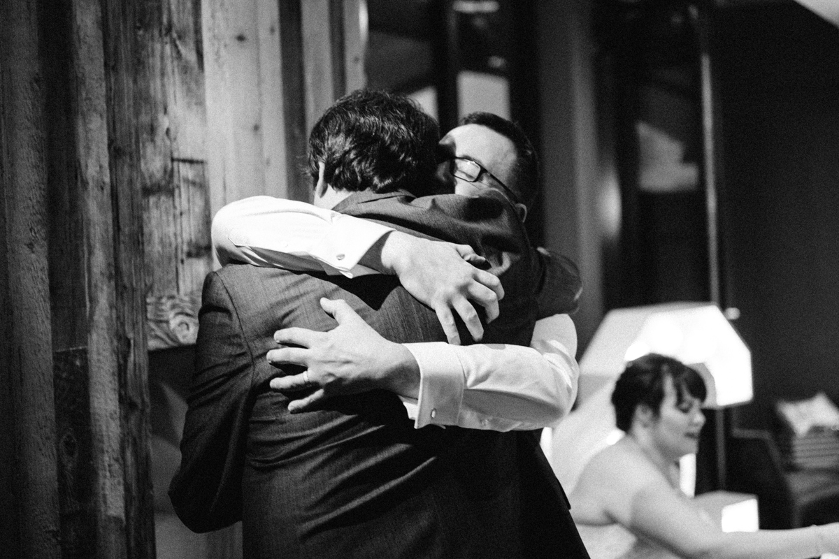 Black and white photograph of the groom stood hugging a suited man with the bride seated in the background