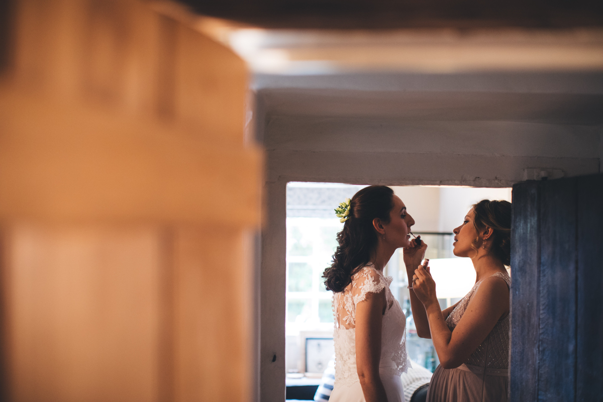 Photograph through a doorway of a bridesmaid putting makeup on the bride