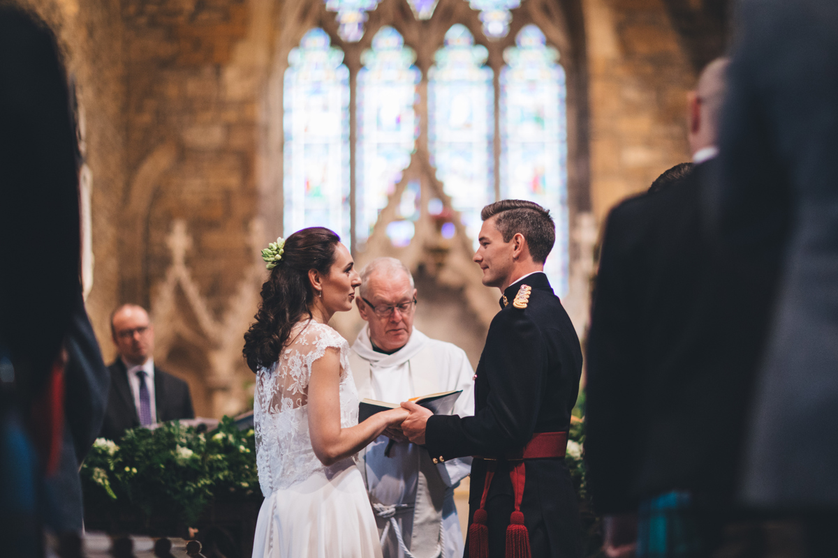 Bride and groom holding hands in front of the priest looking at each other with stained glass windows in the background