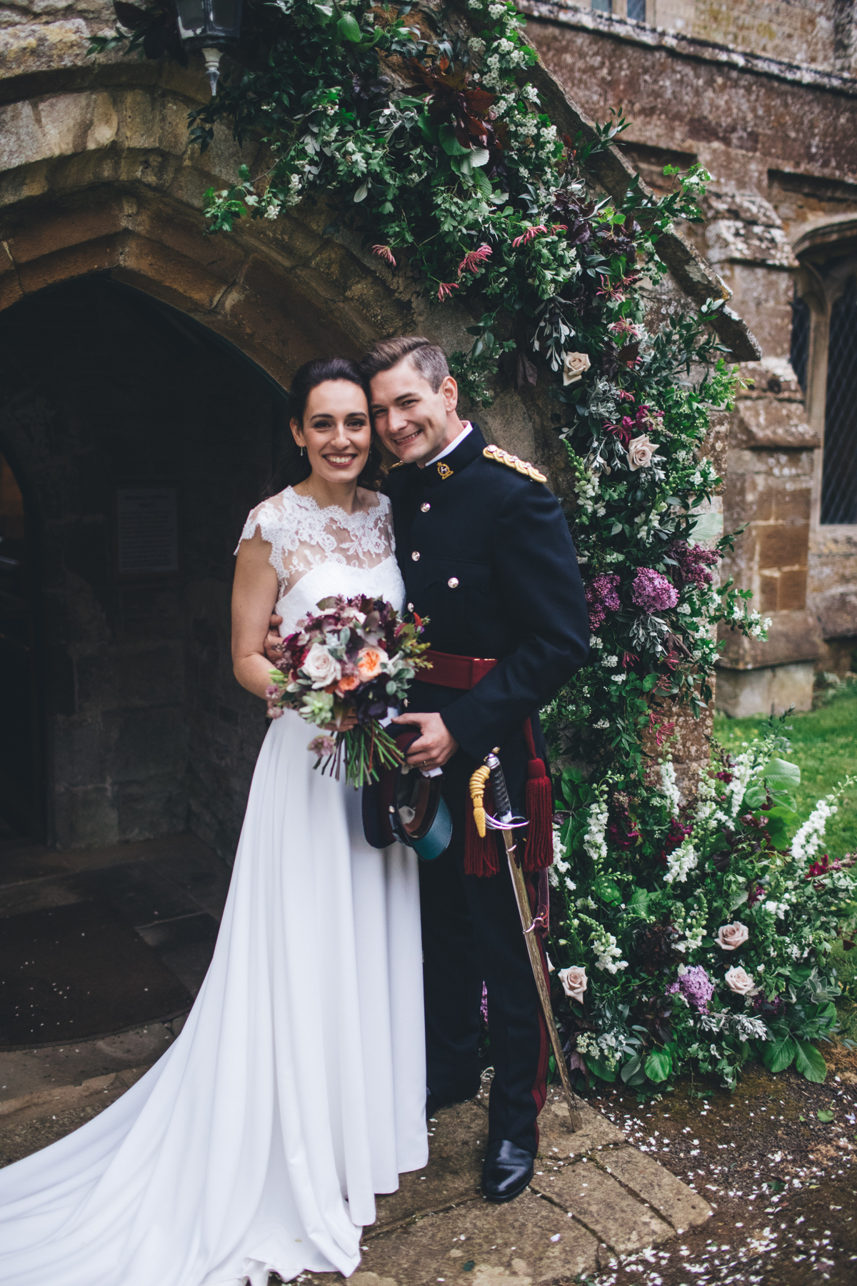 Bride and groom in front of archway at the front of a church with a floral decoration surrounding the right side of the arch