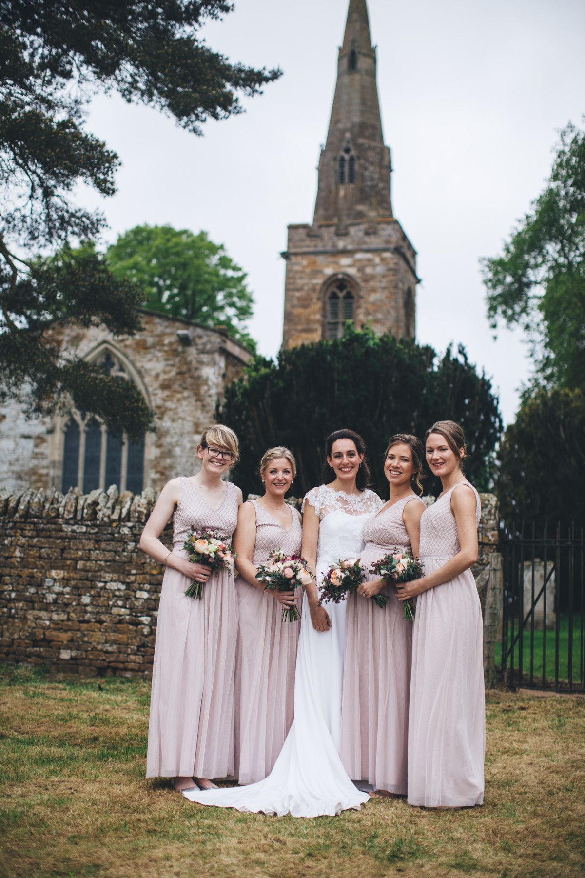 Bride and four bridesmaids stood in front of a stone wall in front of a church spire