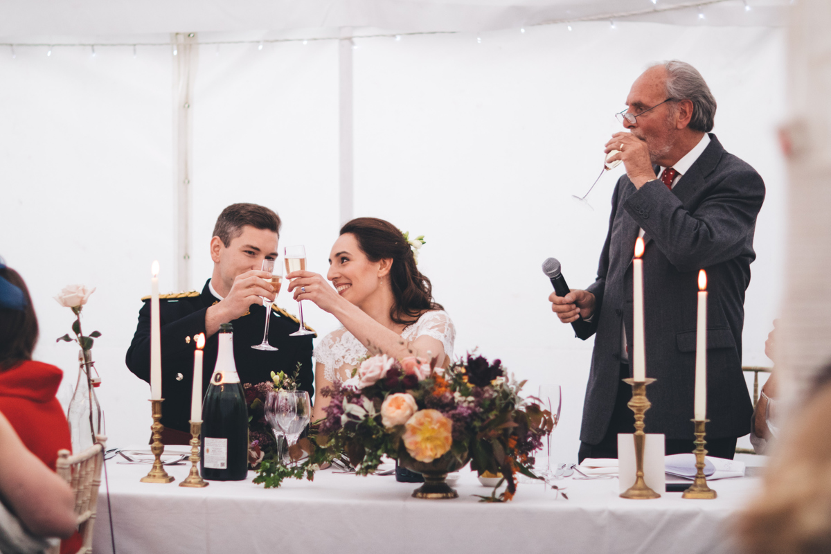 Bride and groom seated clinking champagne glasses whilst the father of the bride is stood with a microphone in one hand and taking a sip of champagne