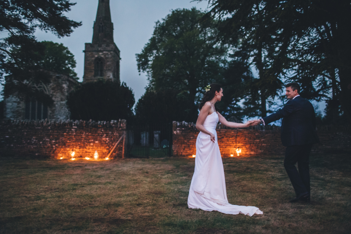 Bride and groom holding outstretched hands in front of a church in the evening
