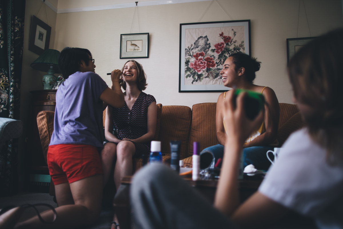 Bride and friends sitting on the sofa with the bride having her makeup done by a woman who is kneeling on the floor in front of her