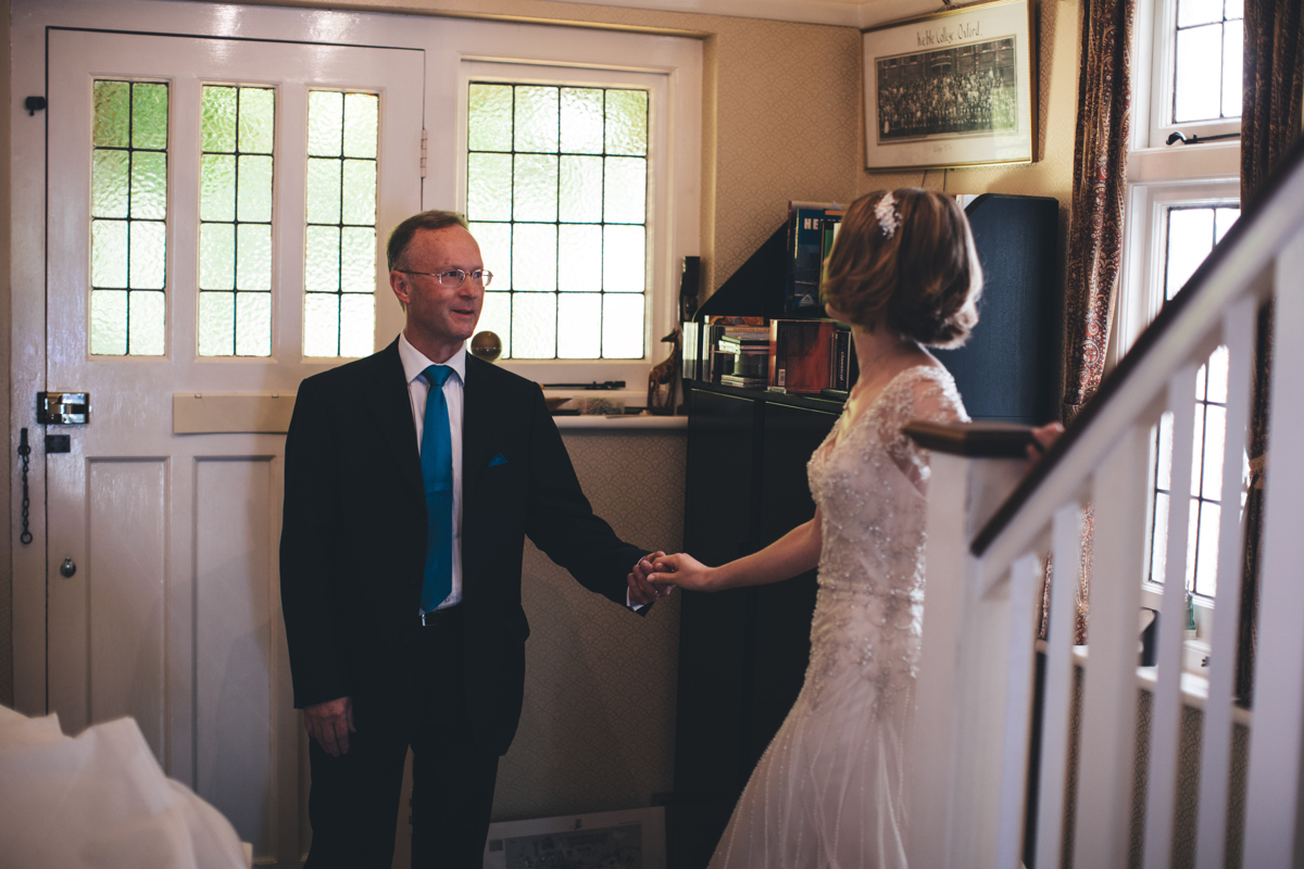 Bride greeting her father at the foot of the stairs at their house. The two of them are holding hands