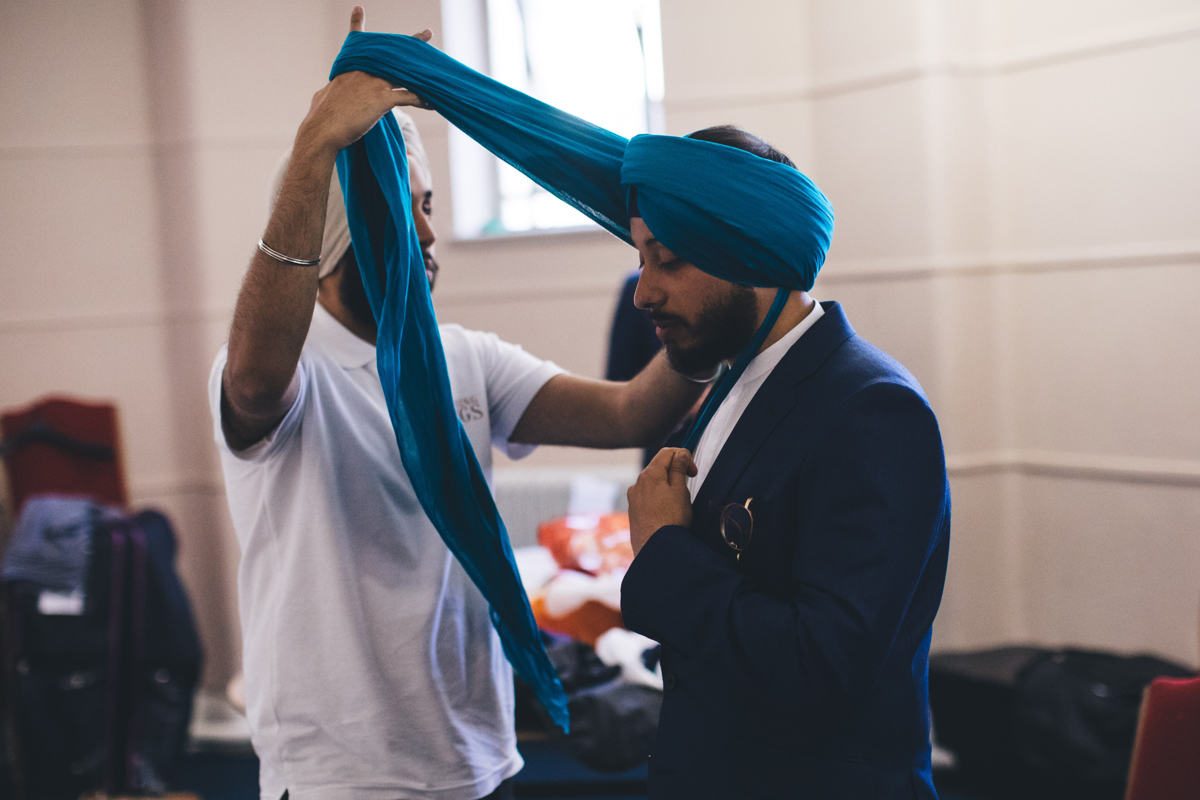 Man in white T-Shirt and cream turban wrapping a blue turban around the head of the Sikh groom