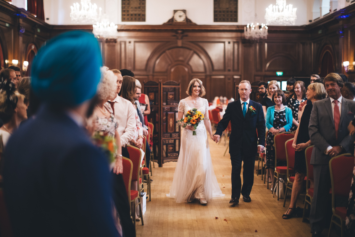 Bride and her father walking down the aisle at Porchester Hall London towards the groom who is pictured from behind and wearing a blue turban