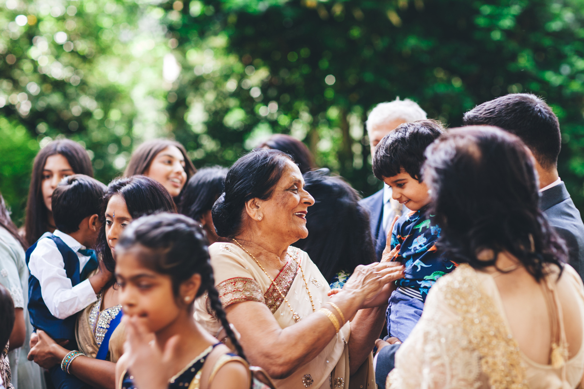 View of an older lady wearing a sari smiling at a young child in a blue outfit in the gardens at Porchester Square London with other members of the wedding party around her