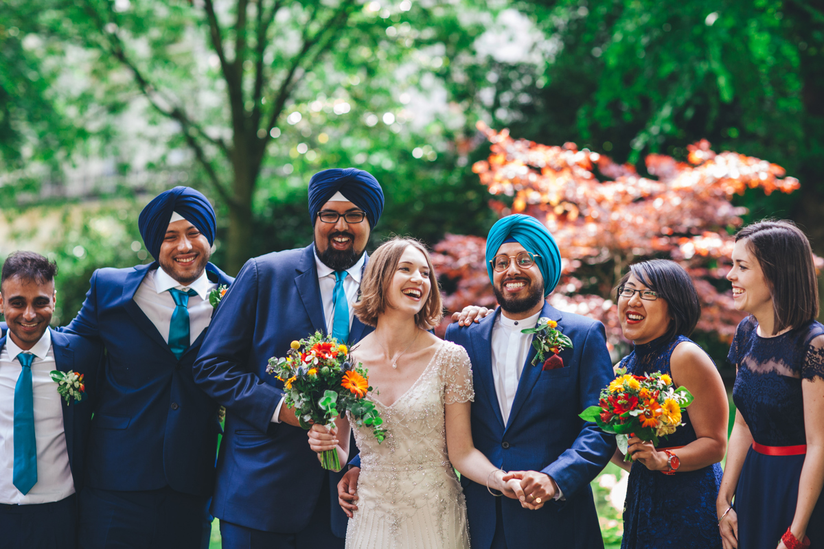 Bride and Groom along with their bridesmaids and groomsmen laughing in the gardens at Porchester Square, London with green and orange trees outof focus in the background