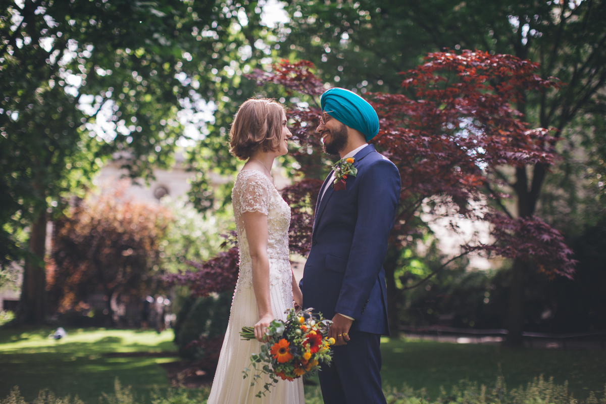 Bride and Groom stood opposite each other, holding one hand with the bride holding a bouquet of flowers in her other hand in the gardens at Porchester Square, London. There are purple Acer trees behind along with other green foliage