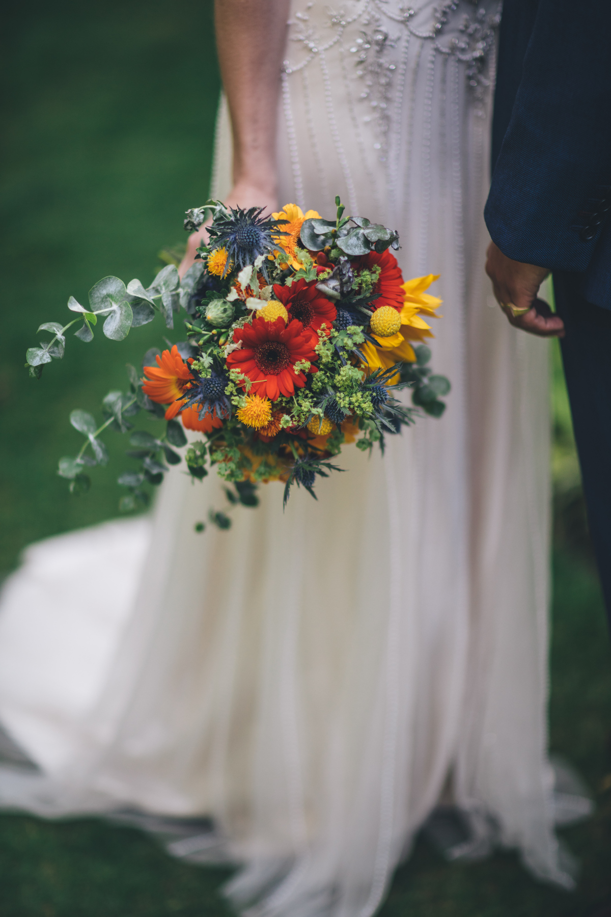 Bride holding a colourful bouquet of orange, yellow and green flowers and foliage, with only the bouquet, the lower half of the brides dress and the grooms arm and hand in the shot