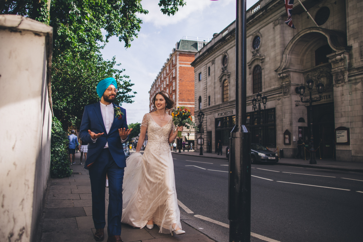 Groom, wearing a suit and a blue turban, with his hands facing upwards walking next to his bride who is holding a bouquet in one hand walking down a street opposite Porchester Hall in central London