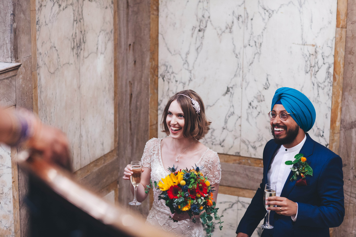 Bride and groom looking upwards towards a person out of shot, both are holding a glass of champagne. There is a marble wall behind them. The bride is holding a bouquet of flowers and the groom is wearing a blue turban and glasses