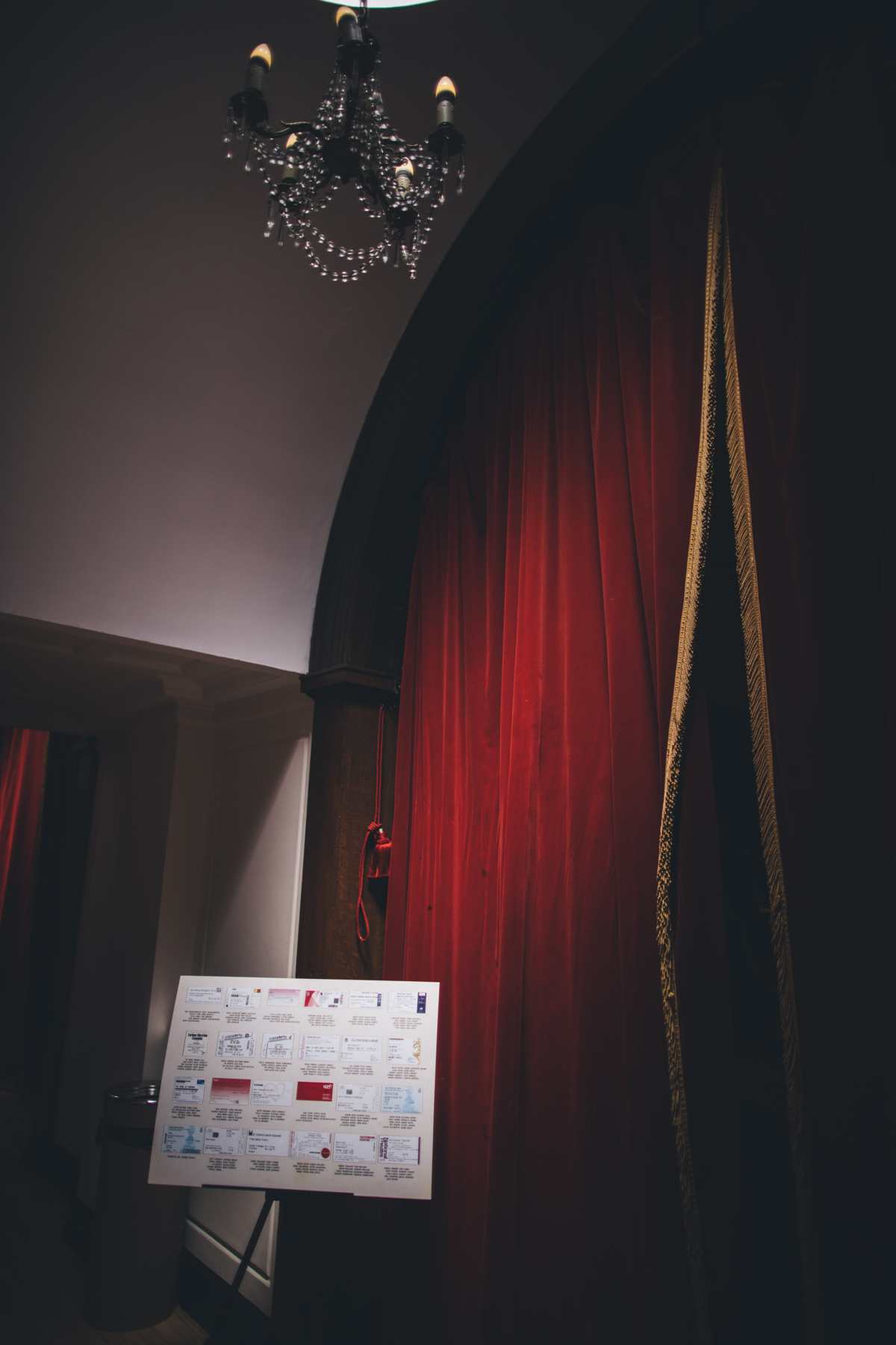 View of a large red velvet curtain with a chandelier above. There is a board in front of the curtain displaying the table plan for the guests at a wedding at Porchester Hall