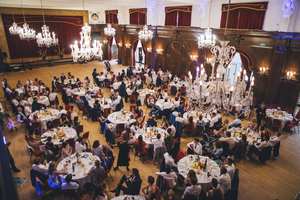 View from the balcony in the Main Hall at Porchester Hall, London, of a wedding reception with many people seated at circular tables. There are a number of chandeliers in the room