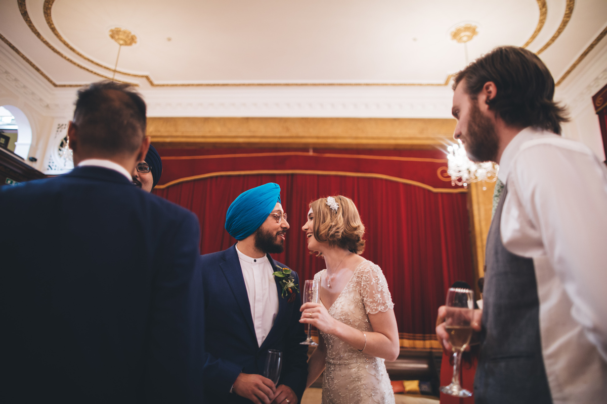 Bride and groom stood close to each other looking into each others eyes in front of a large red velvet curtain in the Main Hall at Porchester Hall, London with two male wedding guests in the foreground facing away from the camera