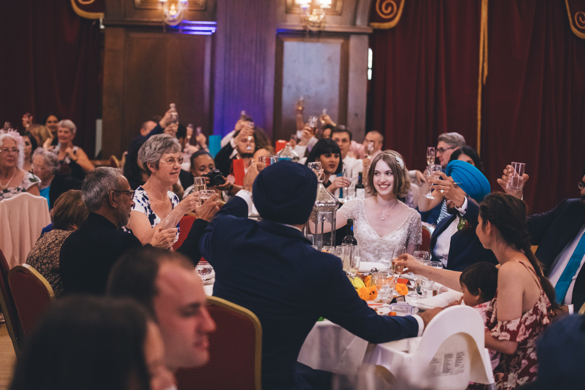 A toast at a wedding reception in the Main Hall in Porchester Hall, London. The wedding party, who are seated, are raising their champagme glasses with the bride facing towards the camera smiling but not looking at the camera